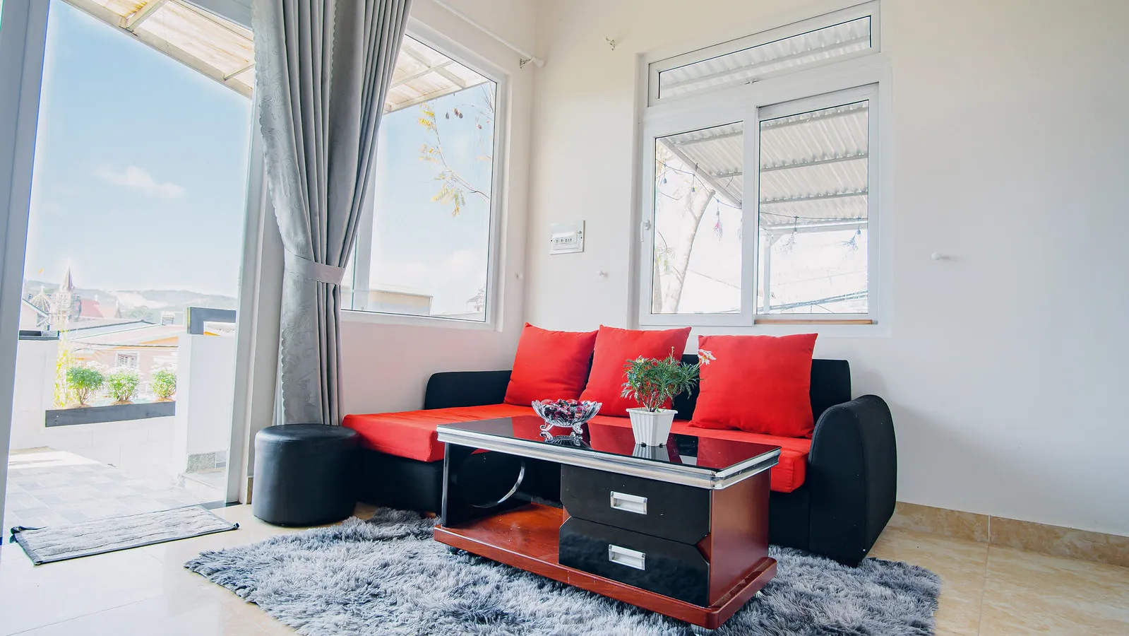 A bright homestay living room with a red sofa and Đà Lạt rooftops through the window.
