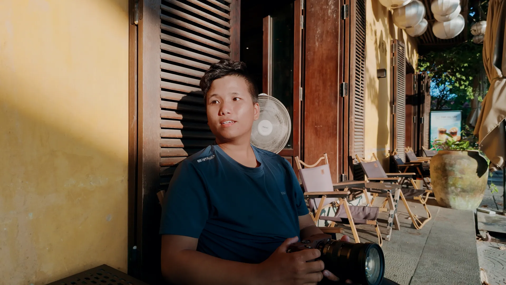 Quốc sitting with his camera outside a sunlit café.