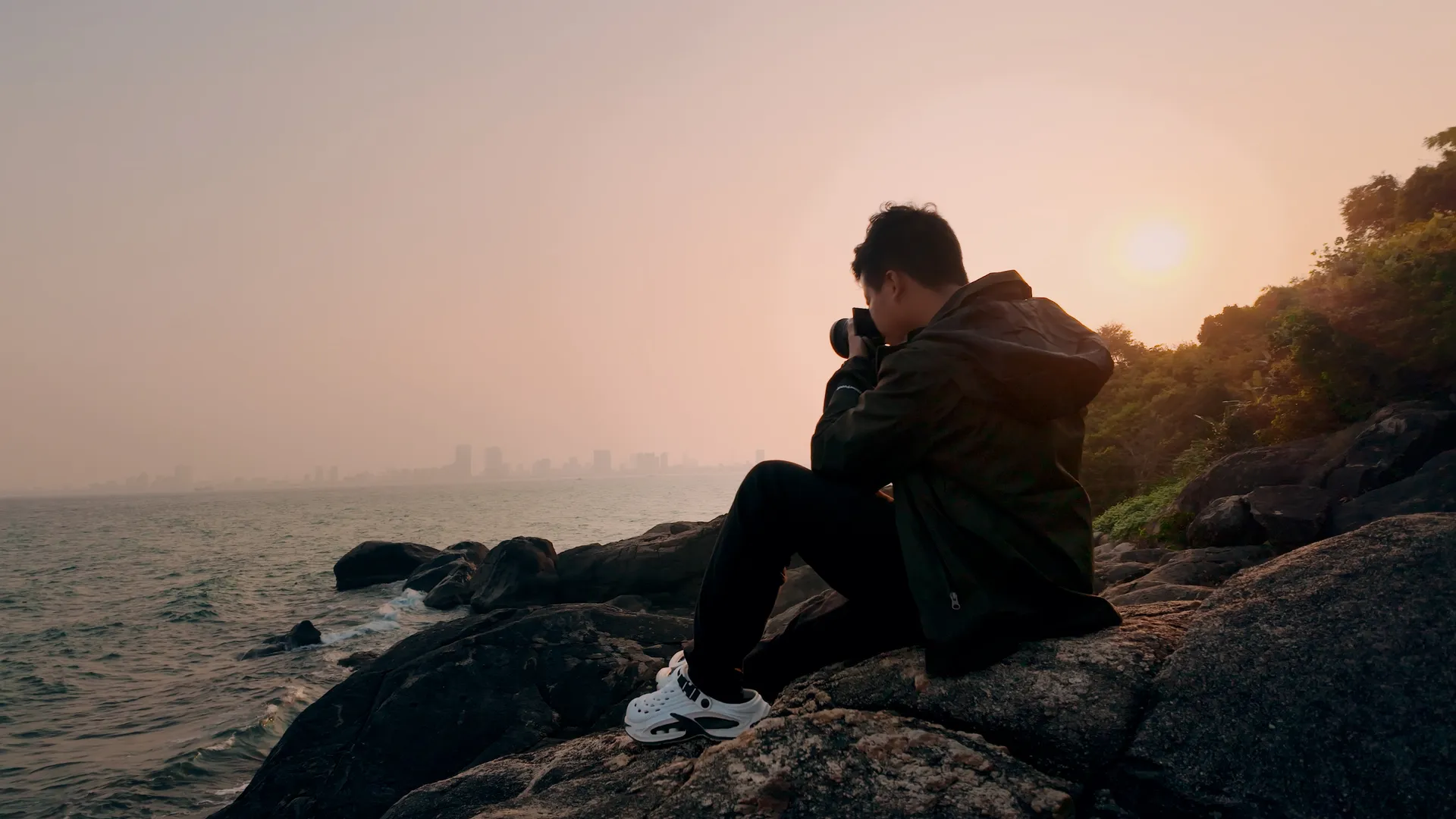 Quốc filming at the coast at sunset, camera raised to his eye.