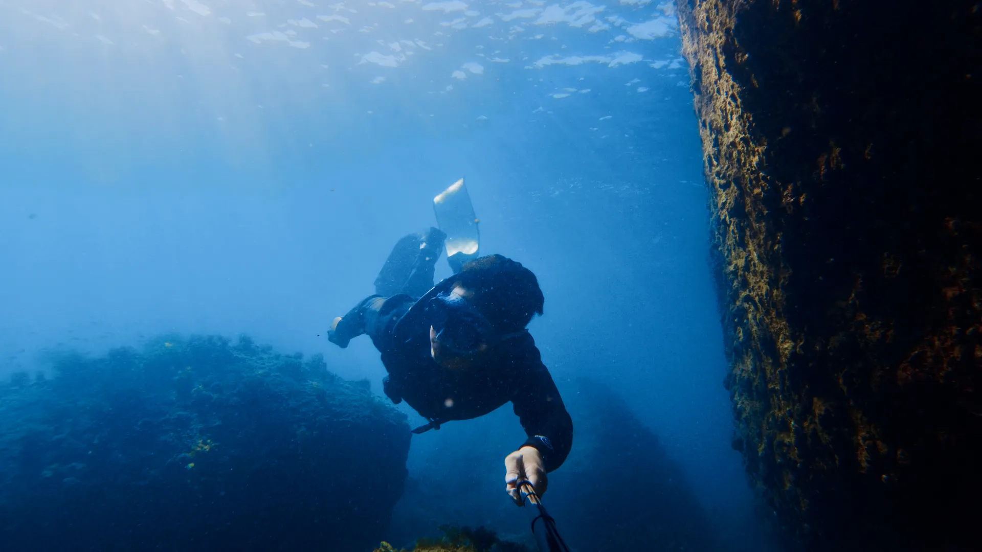A freediver holding mid-water beside a steep rock wall, lit from the surface above.